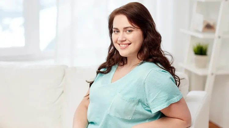 Frau sitzt lächelnd auf Sofa in hellem Raum mit blauem Shirt.