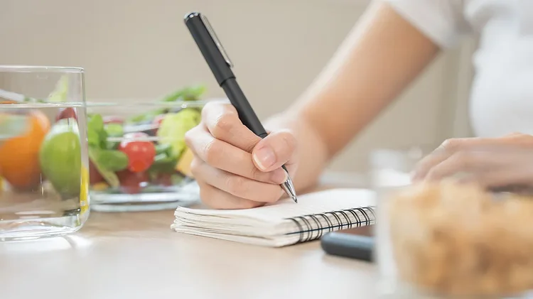 Person schreibt Ernährungsnotizen neben Gemüse und Wasser.