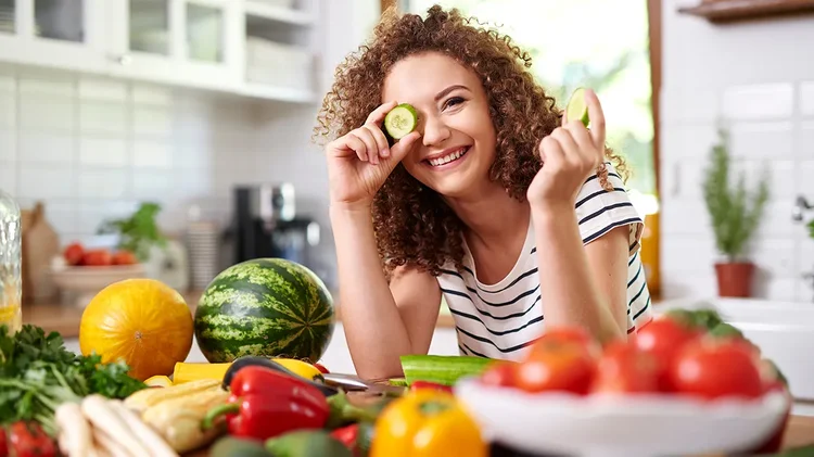 Frau hält Gurkenscheiben vor ihr Gesicht und sitzt zwischen frischem Gemüse.