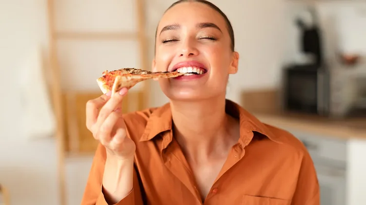 Frau in Küche beißt lachend in ein Stück Pizza.