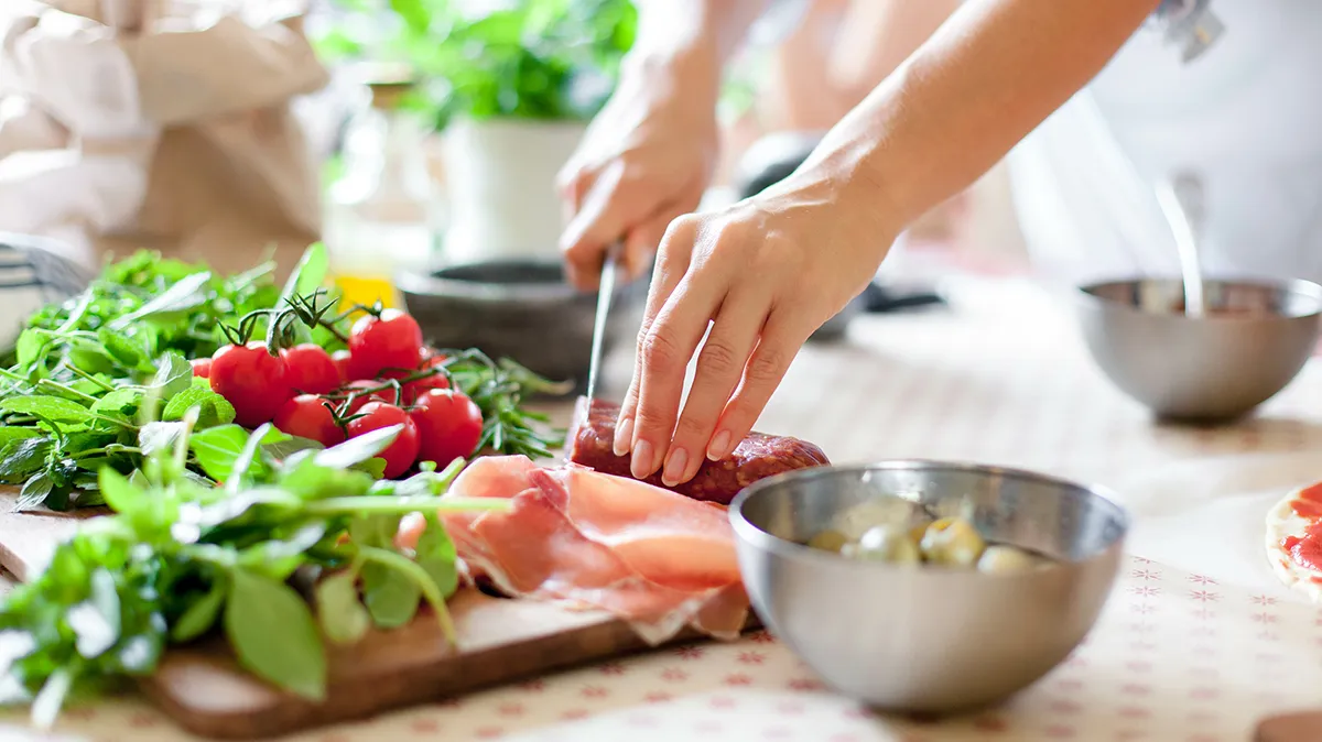 Hände bereiten frische Zutaten wie Tomaten und Schinken auf Schneidebrett zu.