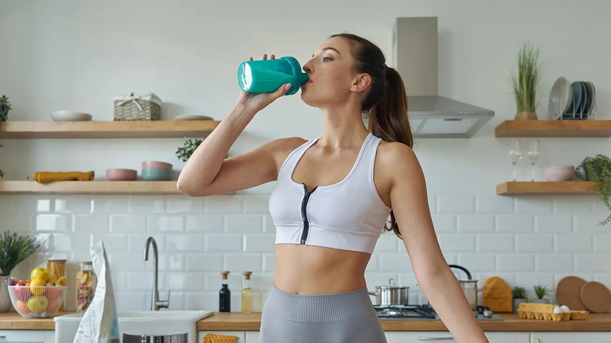Frau in Sportkleidung trinkt Shake in Küche.