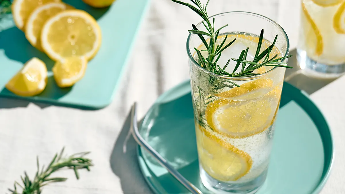 Glas Zitronenwasser mit Rosmarin auf türkisem Teller.