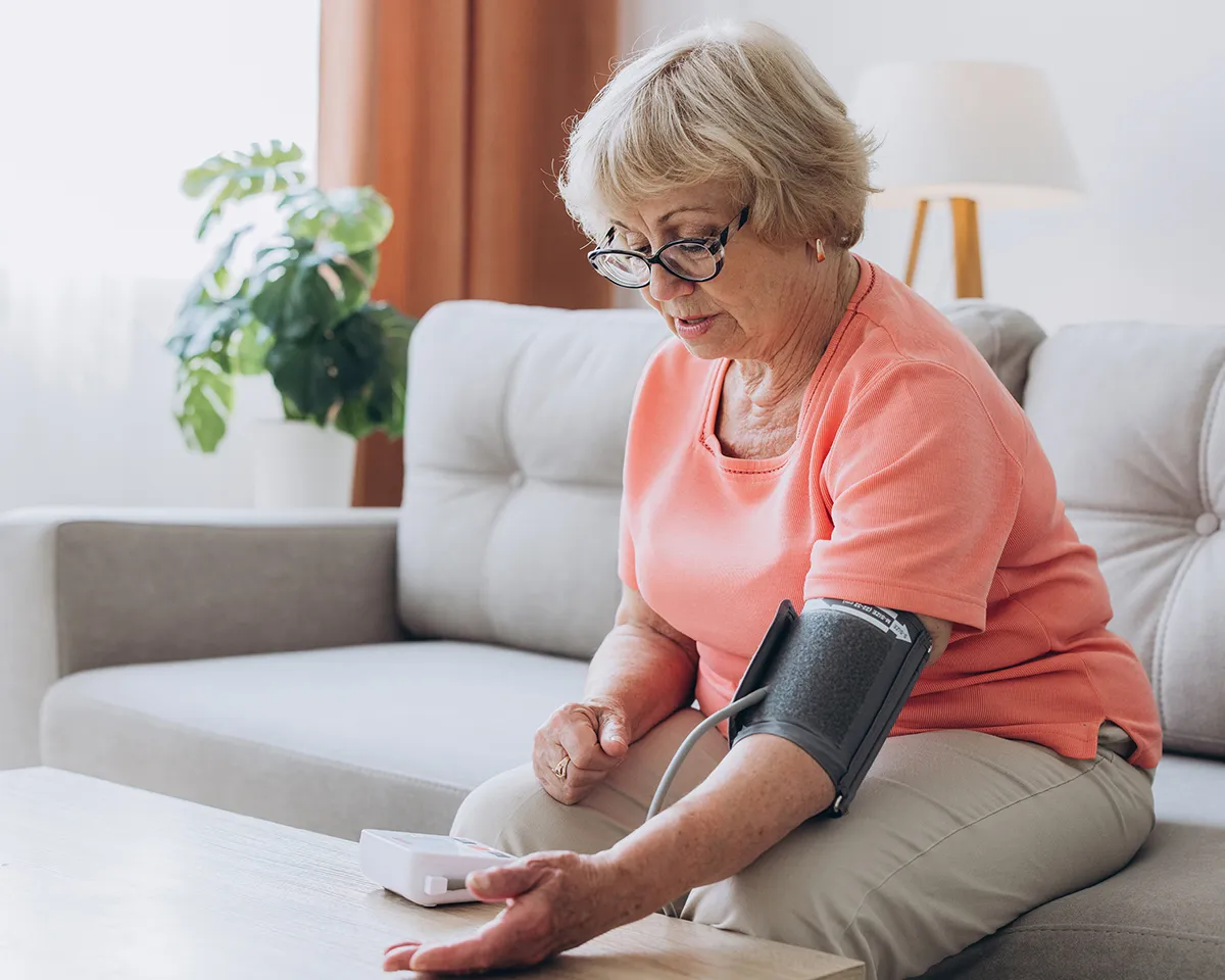 Ältere Frau misst Blutdruck mit Messgerät auf dem Sofa.