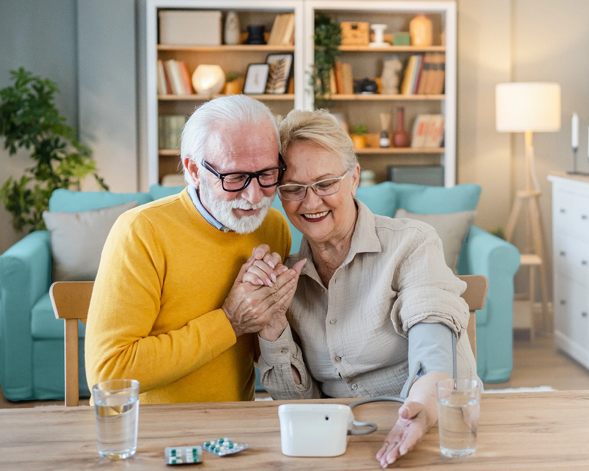 Älteres Paar misst gemeinsam Blutdruck zu Hause und lächelt.