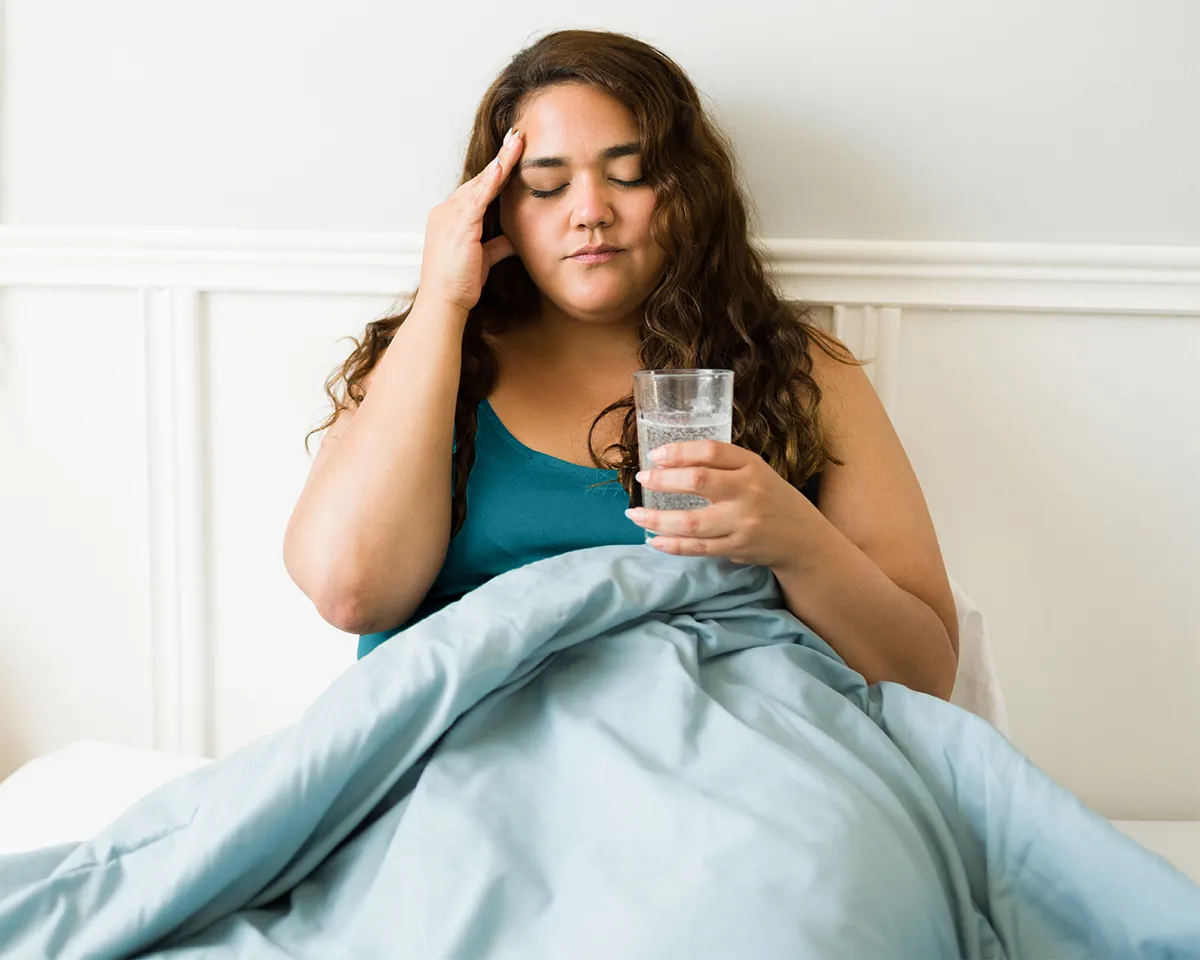 Frau im Bett hält Glas Wasser und wirkt erschöpft.