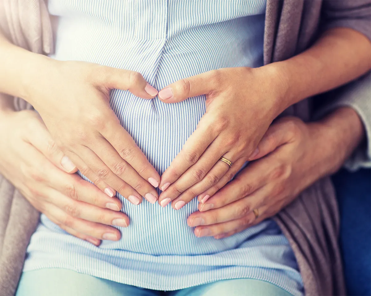 Hände eines Paares formen ein Herz auf einem schwangeren Bauch.