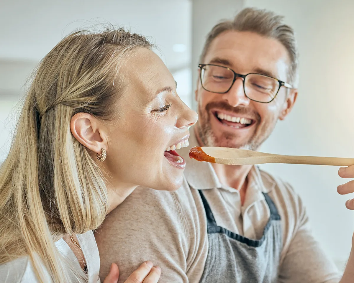 Frau probiert Soße vom Holzlöffel, Mann umarmt sie lachend.