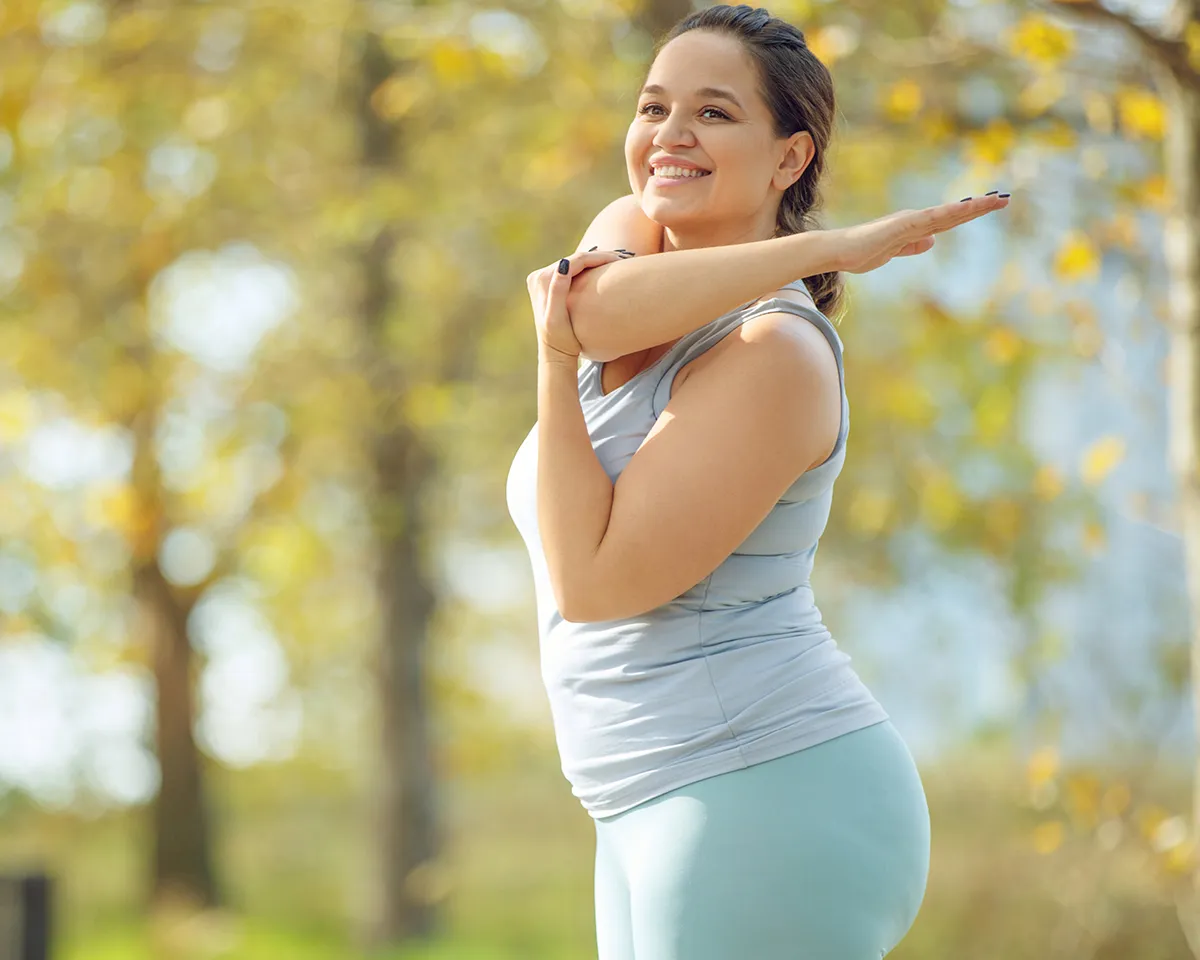 Frau dehnt im Park ihre Arme und lächelt dabei.