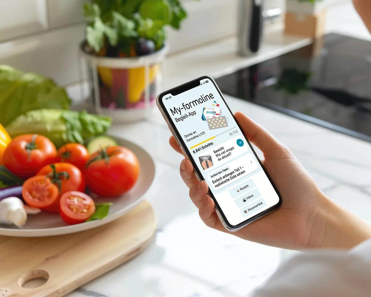 Person hält Smartphone mit geöffneter My-formoline App über einem Tisch mit Gemüse.