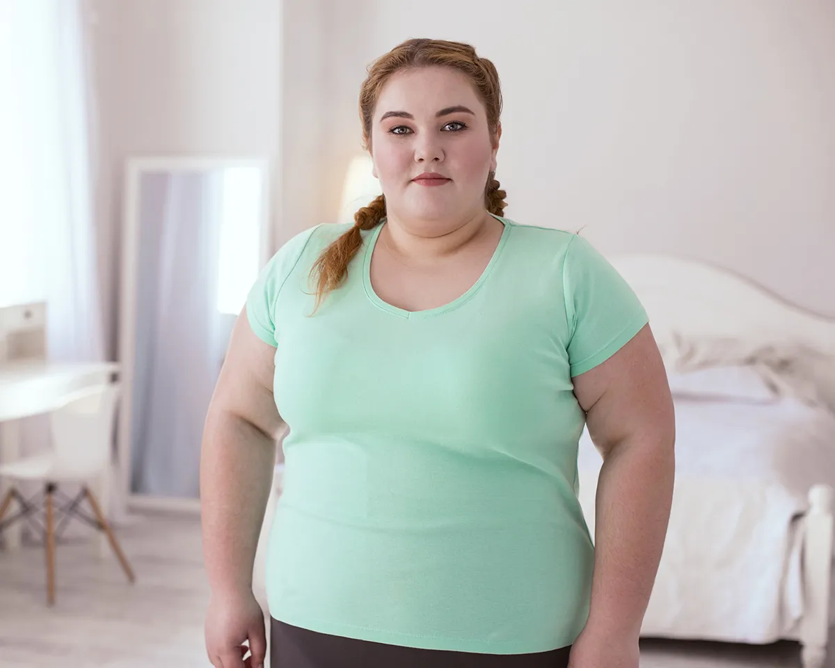 Frau mit Übergewicht steht in hellem Schlafzimmer und blickt in die Kamera.
