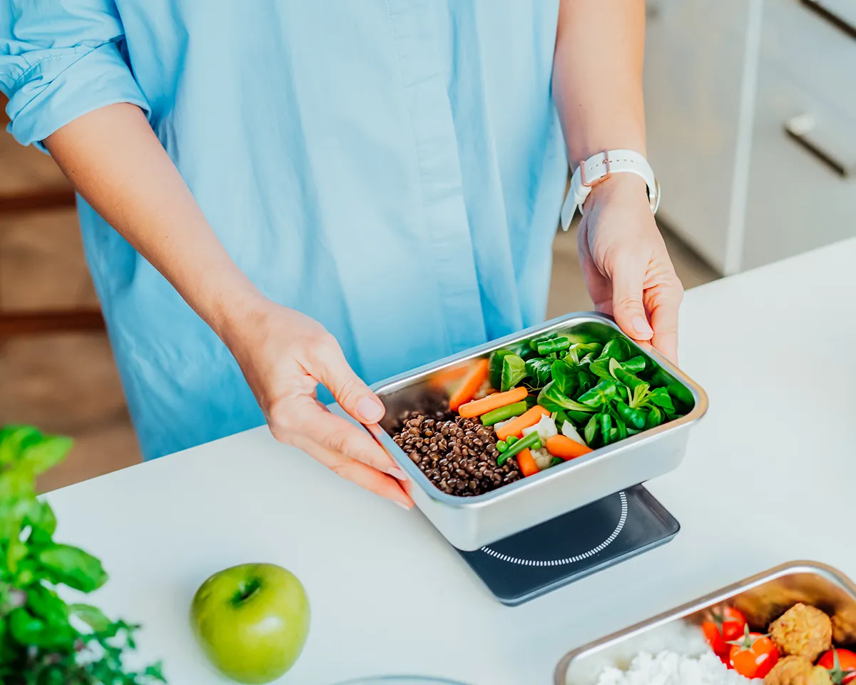 Person wiegt Schale mit Gemüse und Linsen auf einer Küchenwaage ab.