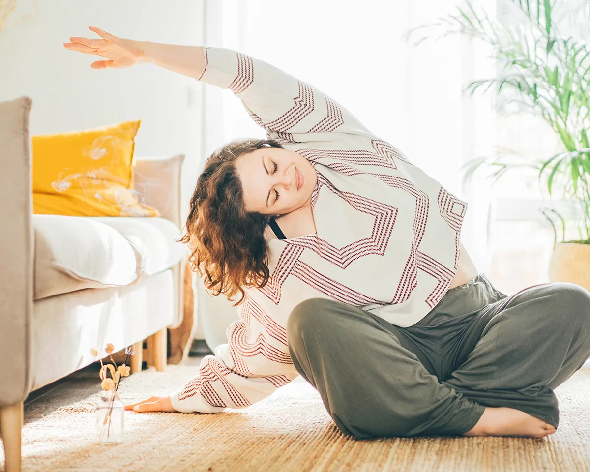Frau macht im Wohnzimmer eine Dehnübung im Sitzen.
